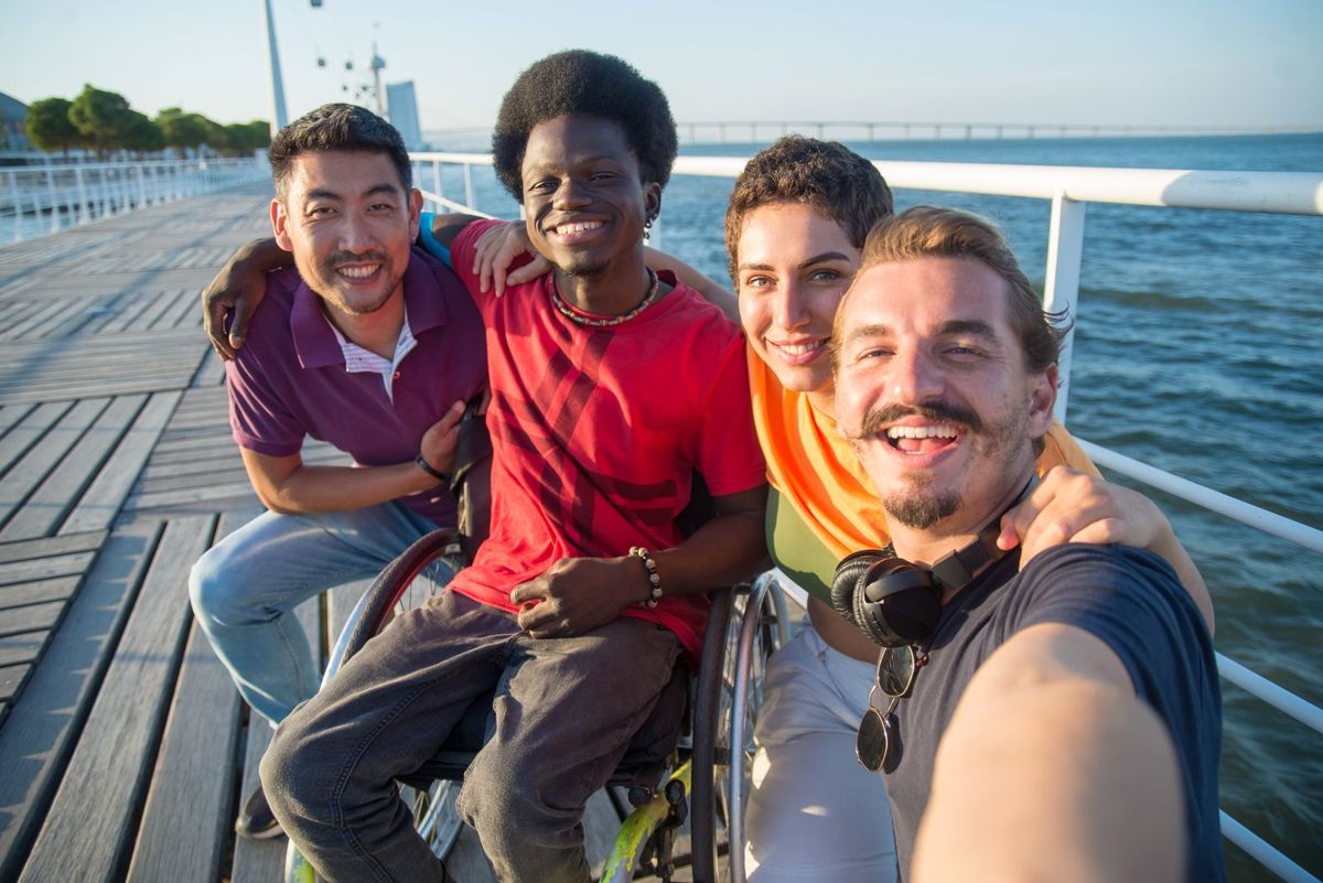 Four smiling young adult friends of different races, one using a wheelchair, take a selfie on a boardwalk